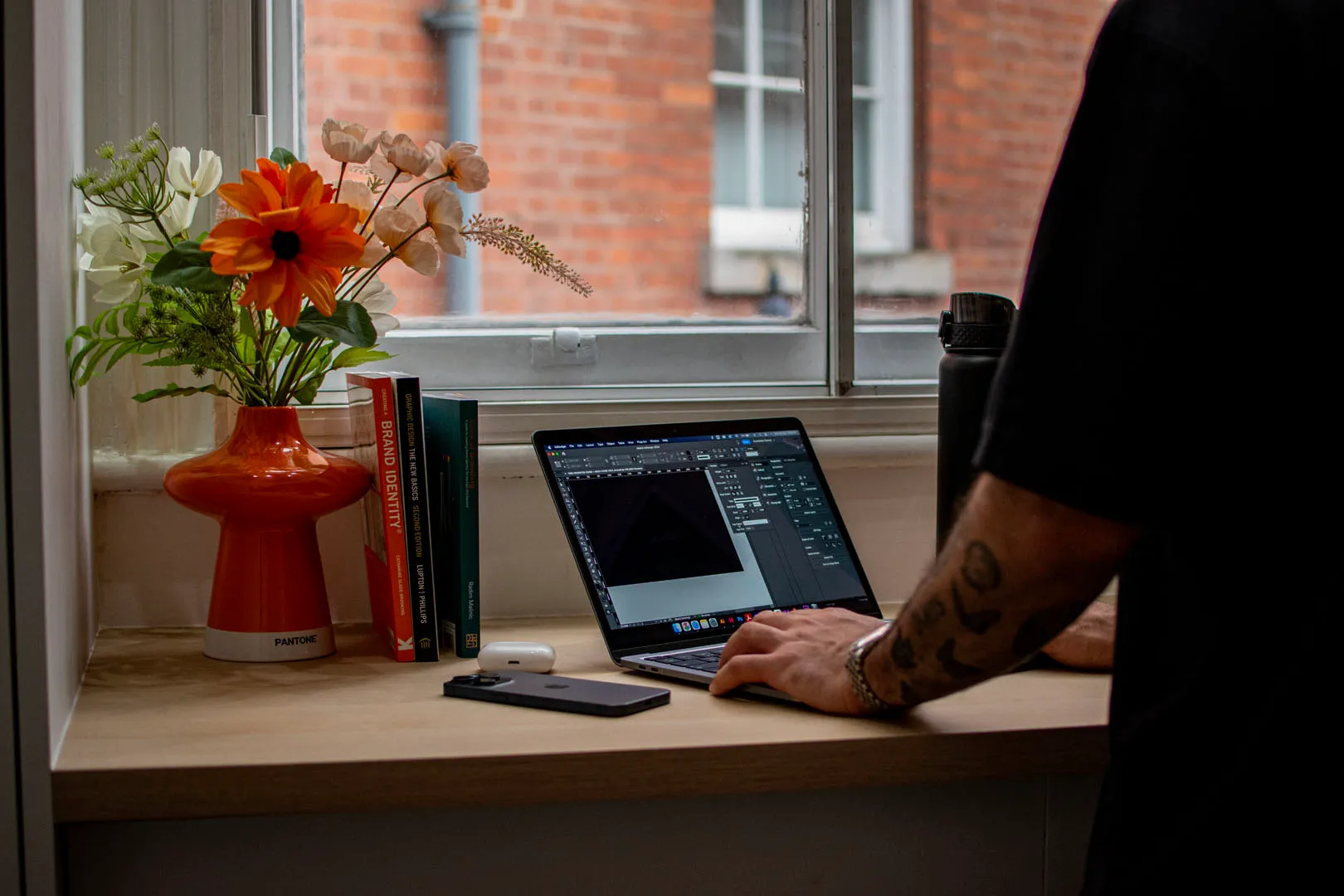 A book titled "Creating a Brand Identity" lies on a wooden desk, accompanied by a laptop and a silver laptop stand. The book's cover features a white background with an orange stripe and black text, while the laptop has a black keyboard wit