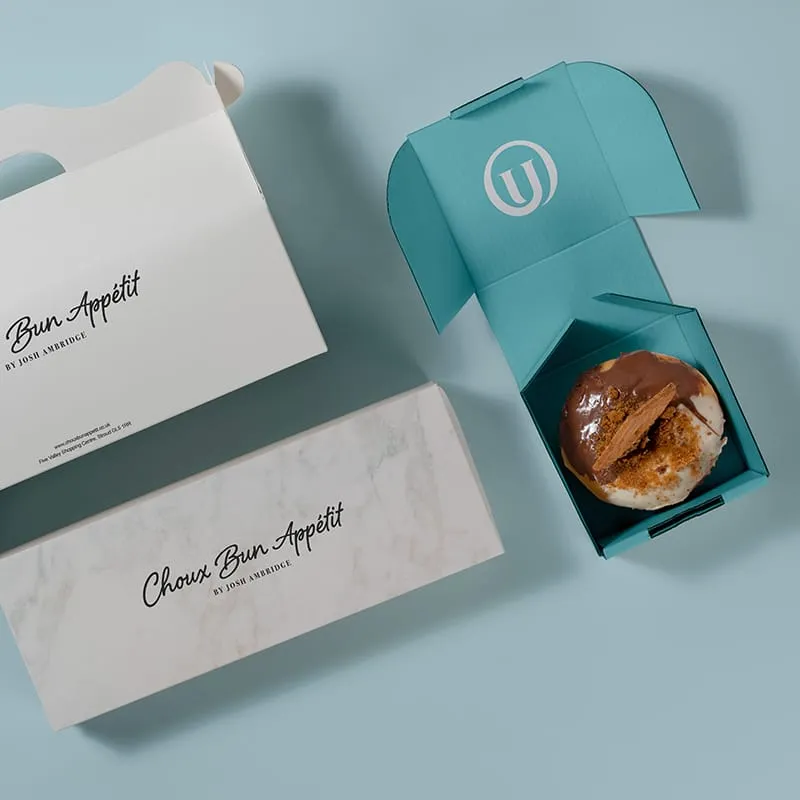 A collection of bakery boxes, one open with a donut inside, on a light blue background. The boxes feature the "Bun Appetit" and "Choux Bun Appetit" logos, with the latter box closed.
