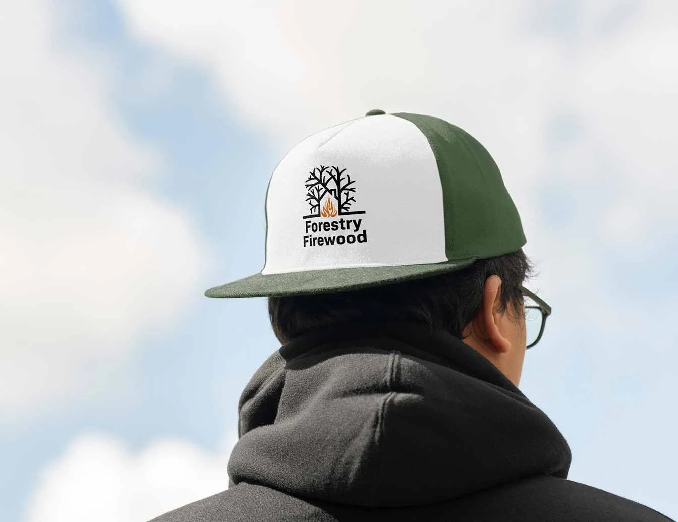 A man wearing a green and white baseball cap with a logo that reads "Forestry Firewood" and a pair of glasses, set against a blue sky with white clouds.